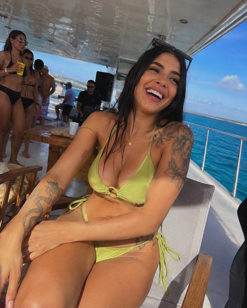 A laughing woman in a lime green bikini sits on a boat deck, with other people in swimwear visible in the background on a sunny day at sea.