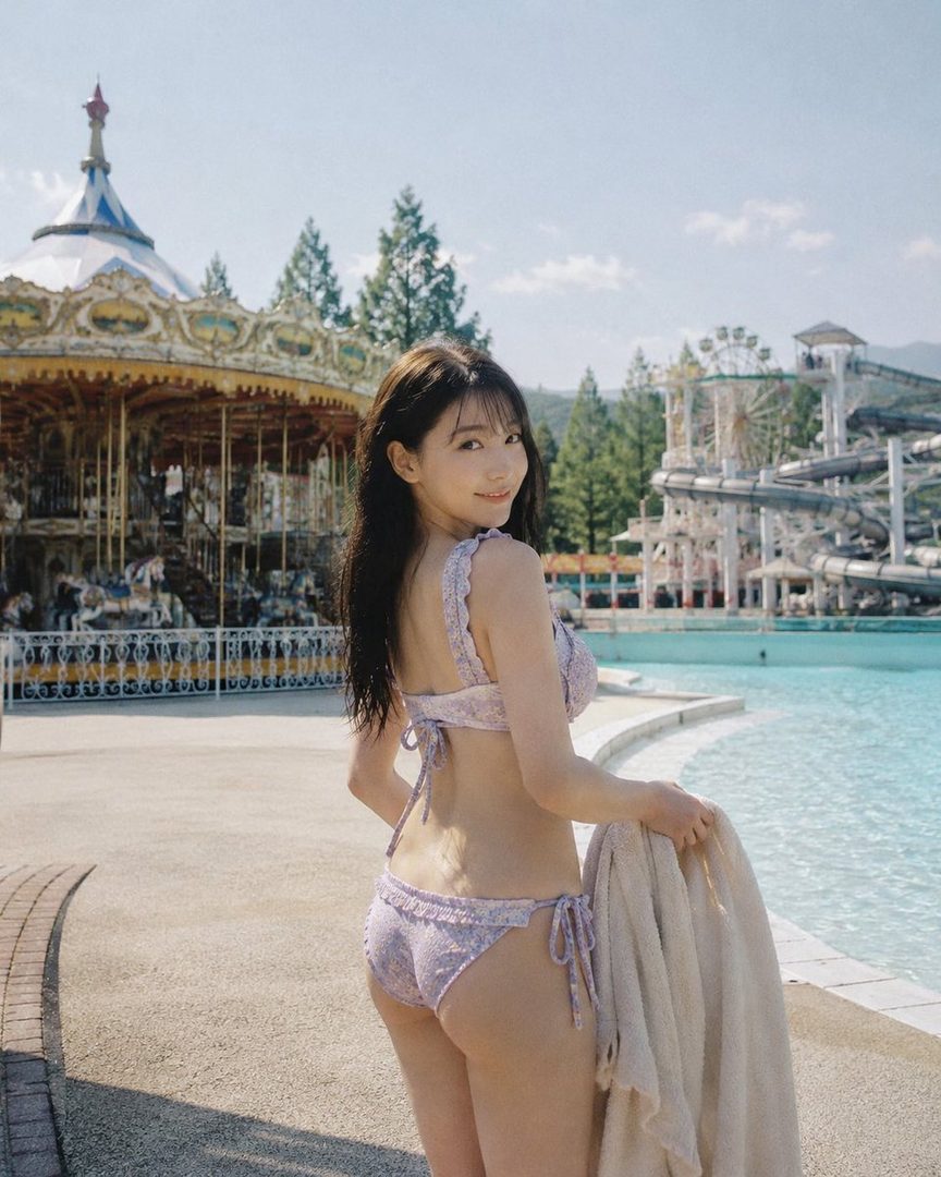 A young woman in a purple bikini poses by a swimming pool at an amusement park, holding a towel.