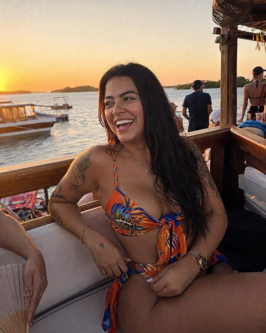 A smiling woman in a colorful bikini poses on a boat at sunset.