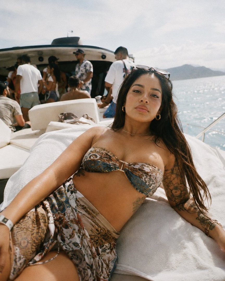 A woman in a bikini and sarong lies on a yacht deck, with other people in the background and the ocean beyond.