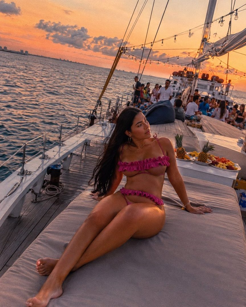 A woman in a pink bikini sits on the deck of a sailboat at sunset, with the ocean and a distant city skyline in the background.