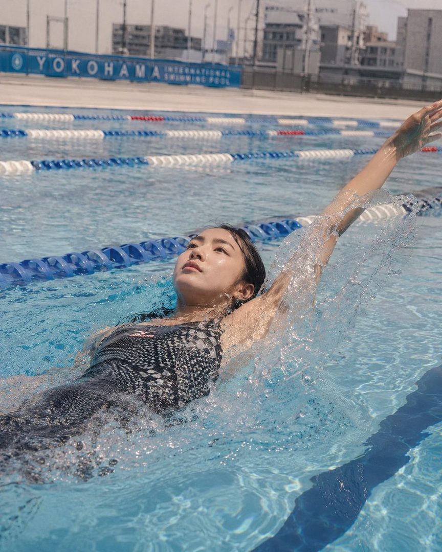 A young East Asian woman in a patterned swimsuit swims in a pool, with lane markers and the Yokohama International Swimming Pool sign visible in the background.