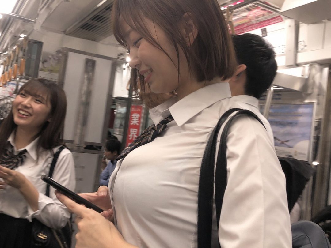 Two young women in school uniforms smiling on a train, one looking at her phone.