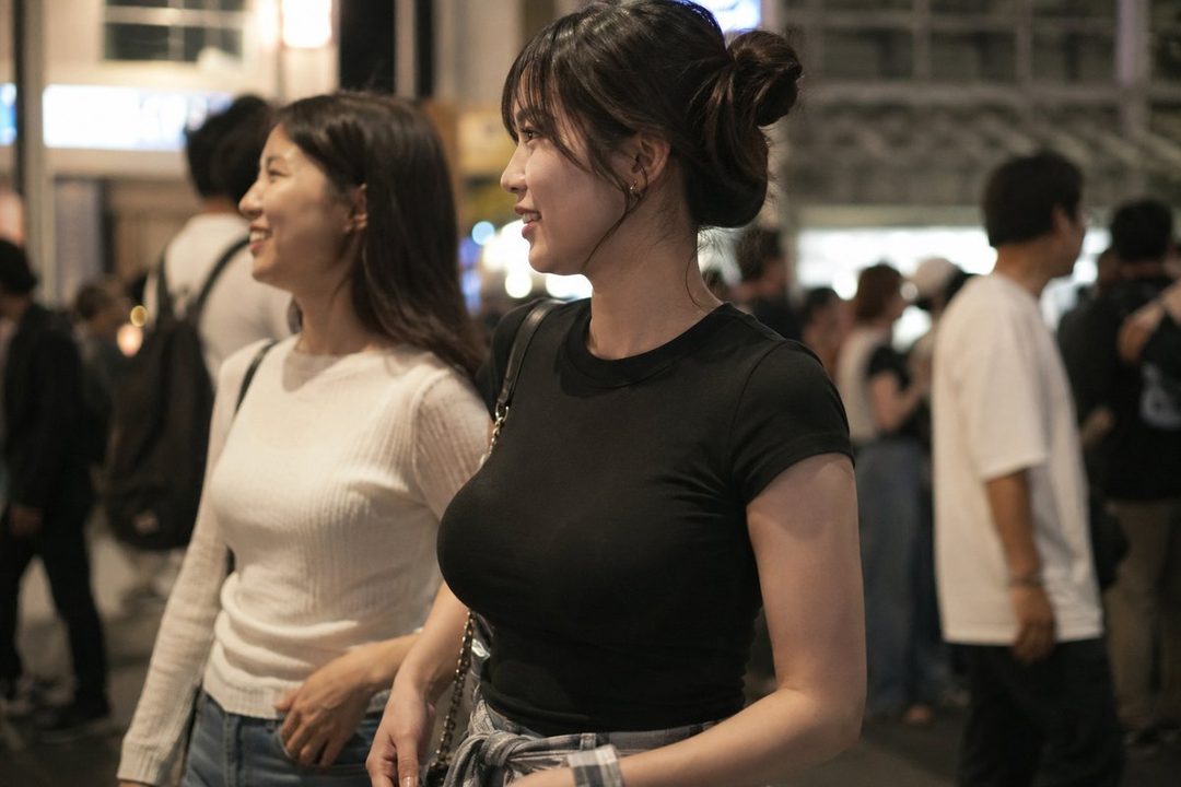 Two young East Asian women walking on a city street at night, one in a black t-shirt and the other in a white long-sleeved shirt.
