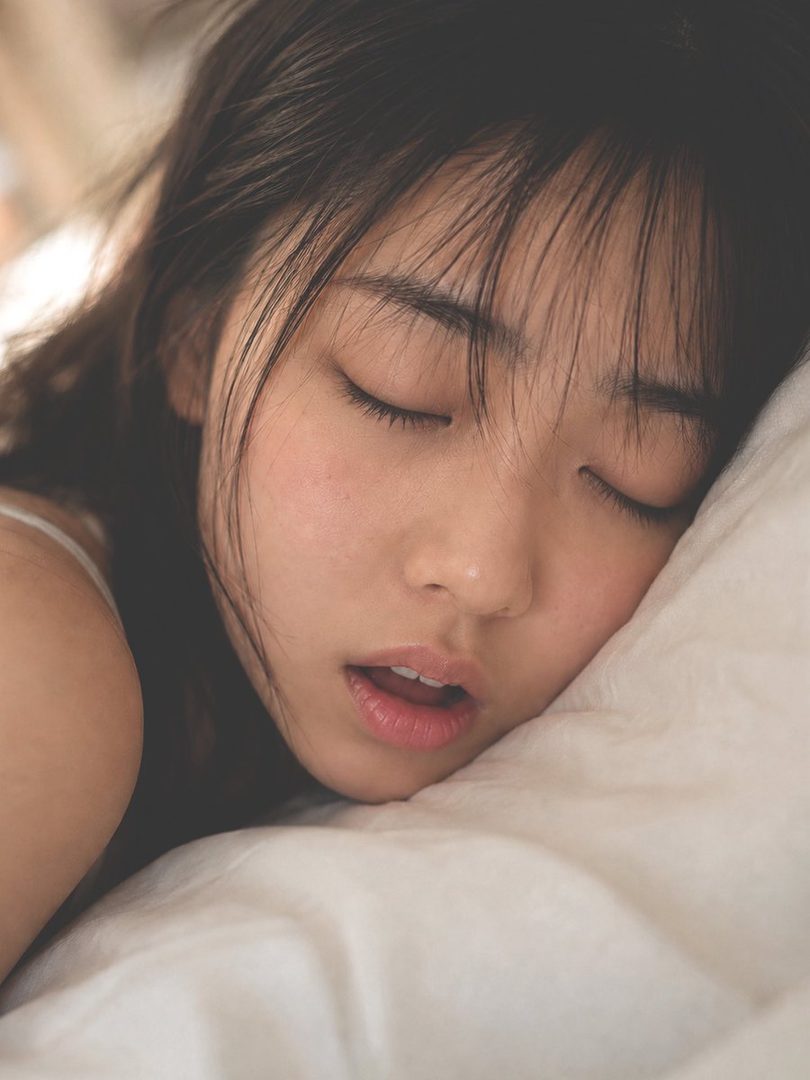 A close-up portrait of a young East Asian woman sleeping peacefully on a white pillow, with her eyes closed and mouth slightly open.