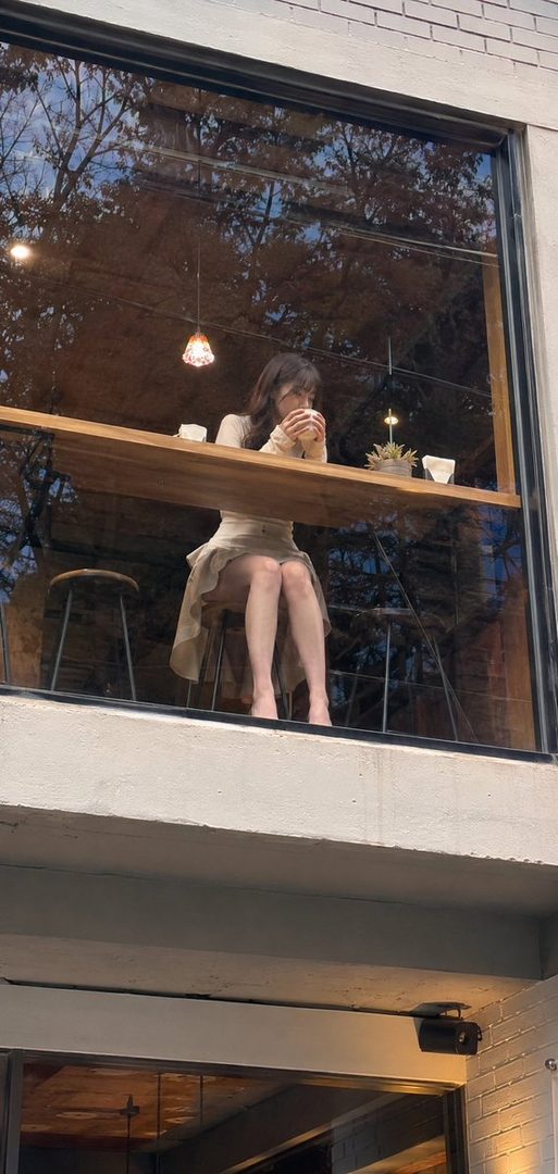 A woman with long brown hair sits at a window counter in a cafe, holding a cup and looking out.