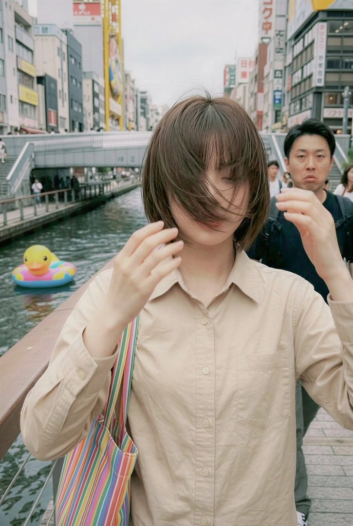 A young woman with short brown hair covering her face stands on a bridge in a city, holding a colorful tote bag. A man with a surprised expression is visible behind her, and a rubber duck float is in the canal below.