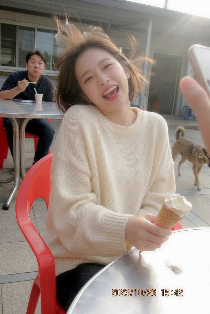 A young woman with short brown hair, wearing a cream-colored sweater, laughs joyfully while holding an ice cream cone. A man is visible in the background, and a dog walks by.