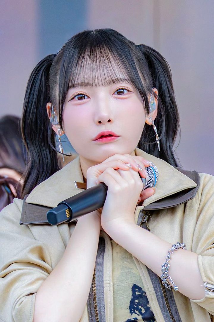 A young East Asian woman with black hair in twintails, wearing a beige jacket, holds a microphone and looks towards the camera with a cute expression.