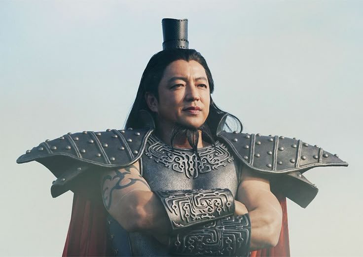 A man in historical armor with long black hair and a goatee, posing with his arms crossed against a clear sky.