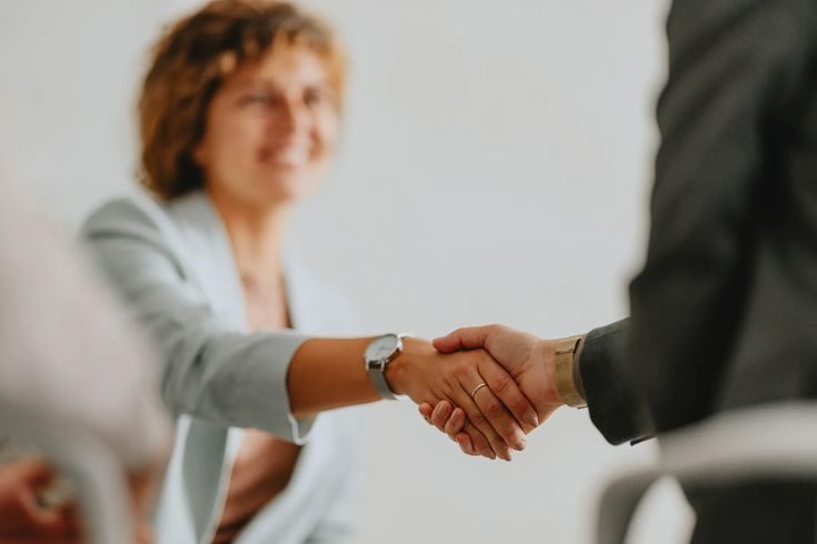 A woman in a light blue blazer and a man in a dark suit shake hands in a professional setting.