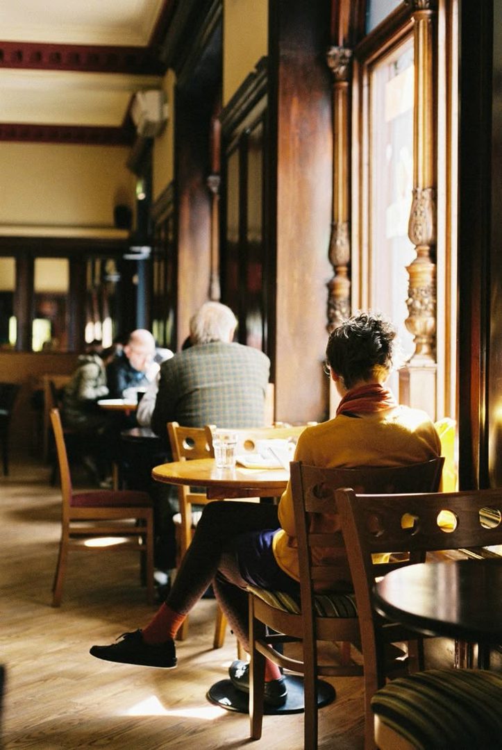 A person sits at a table in a cafe, looking out a window, with other patrons in the background.