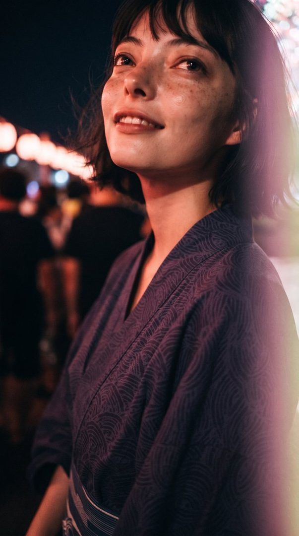 A young woman with short dark hair and freckles smiles gently, wearing a traditional Japanese yukata at a night festival.