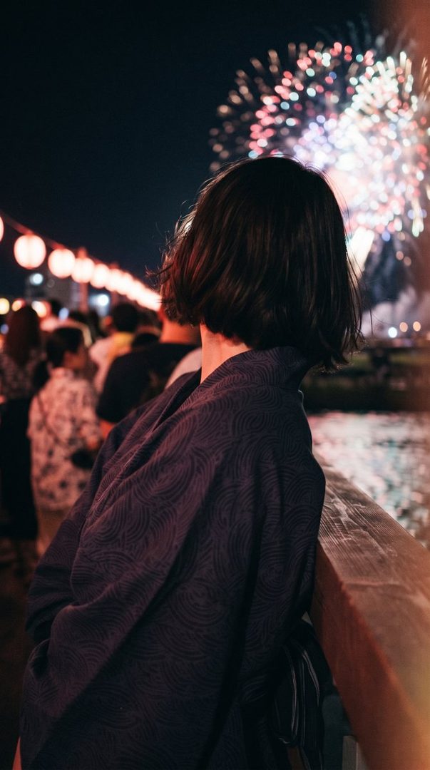 A person in a yukata watches fireworks at a night summer festival.