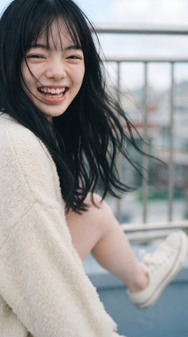 A young East Asian woman with long black hair smiles brightly, wearing a cream-colored sweater and white sneakers, outdoors.