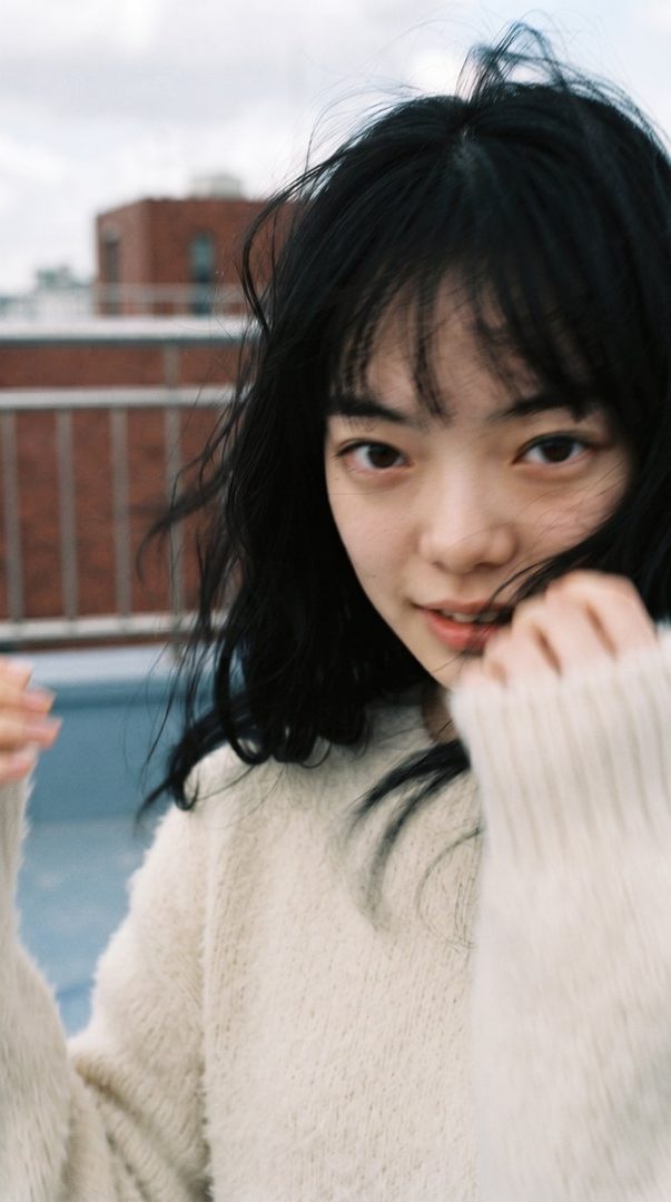A young East Asian woman with long, dark, messy hair and bangs, wearing a cream-colored knit sweater, looks towards the camera with a soft expression. She is outdoors on a rooftop with buildings in the background.
