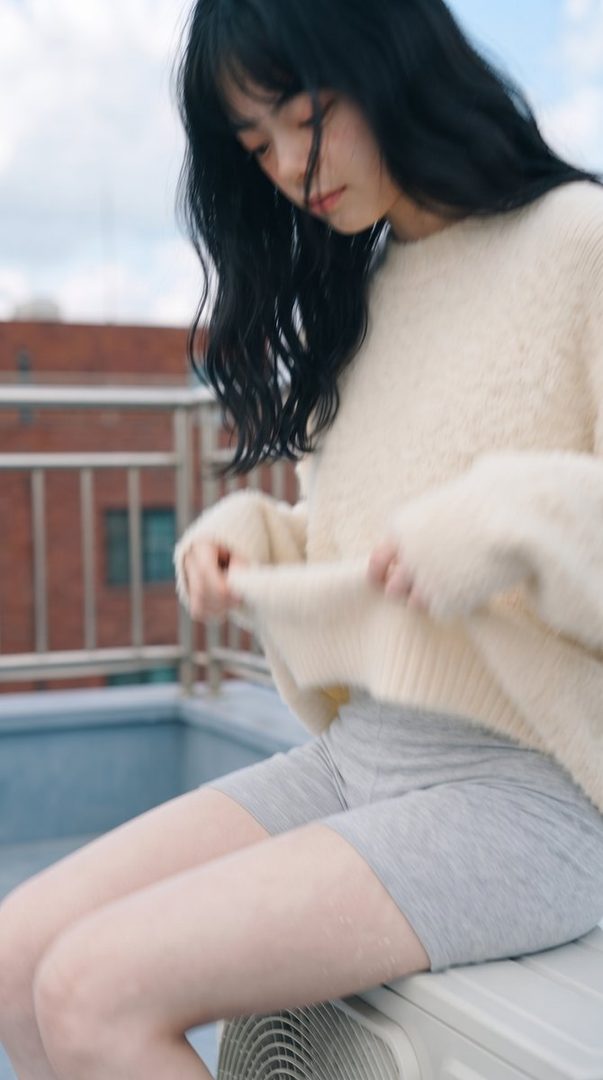 A young East Asian woman with long black hair, wearing a fluffy cream sweater and grey shorts, sits outdoors on an air conditioning unit, looking down.