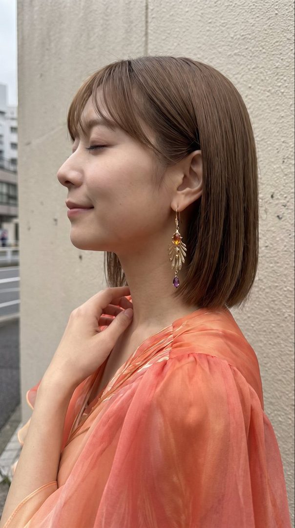 A woman with short brown hair and bangs, wearing a flowing orange blouse and large dangling earrings, poses with her eyes closed and a slight smile.