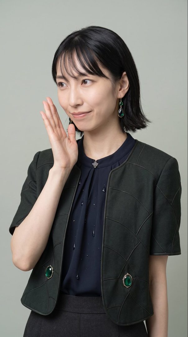 A Japanese woman with short black hair and bangs, wearing a dark blue blouse and a dark green jacket with green jewel accents, poses indoors with her hand near her face.