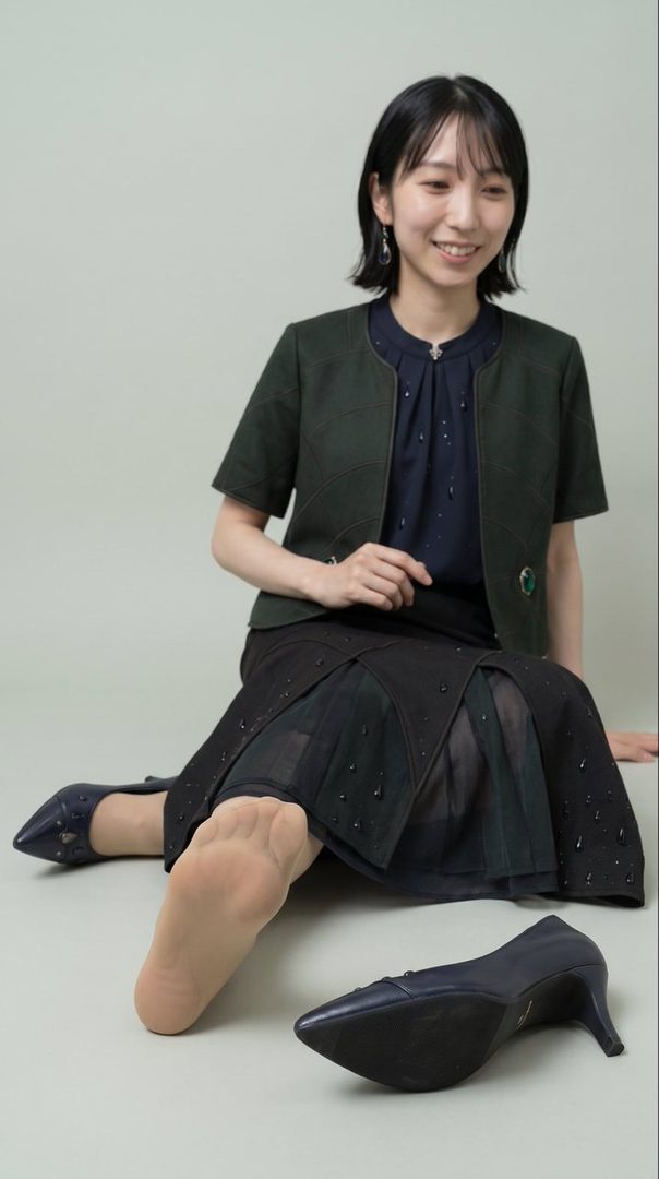 A young East Asian woman with short black hair and bangs, wearing a dark green jacket over a navy blue blouse and a black skirt, sits on the floor with one leg extended, showing her foot in a nude stocking and a navy blue high-heeled shoe nearby.