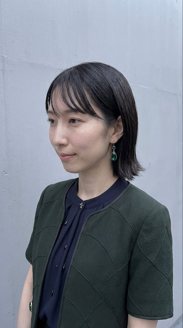 A woman with short black hair and bangs, wearing a dark green jacket over a navy blue blouse and green teardrop earrings, poses outdoors against a gray wall.