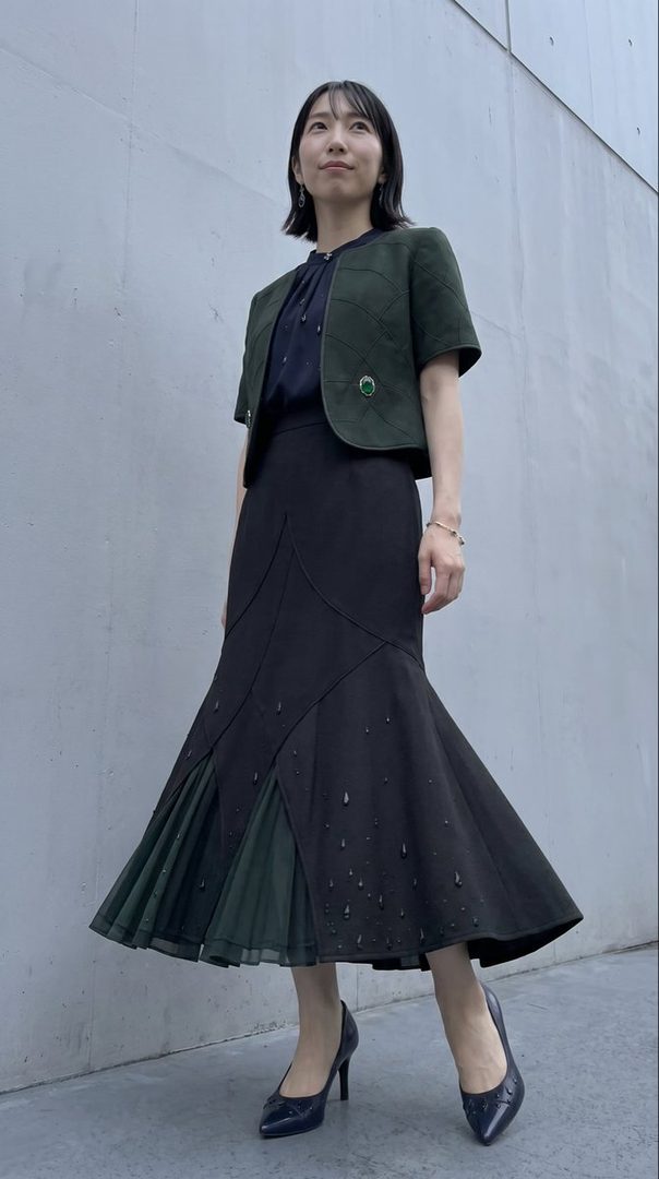 A woman in a dark green jacket and a black fishtail skirt stands against a gray wall.