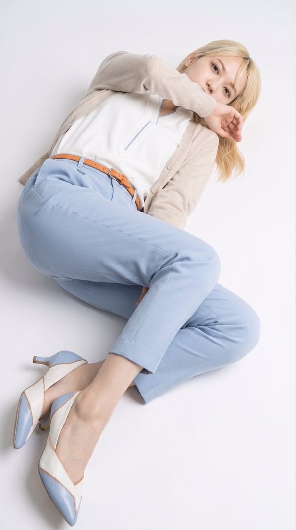 A young woman with blonde hair, wearing a light cardigan over a white top and blue trousers, poses elegantly on a white background.