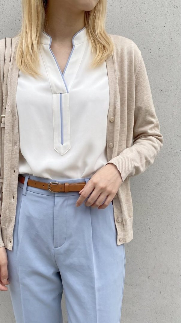 A woman with blonde hair wears a white blouse with light blue trim, a beige cardigan, light blue trousers, and a brown belt, standing against a gray wall.