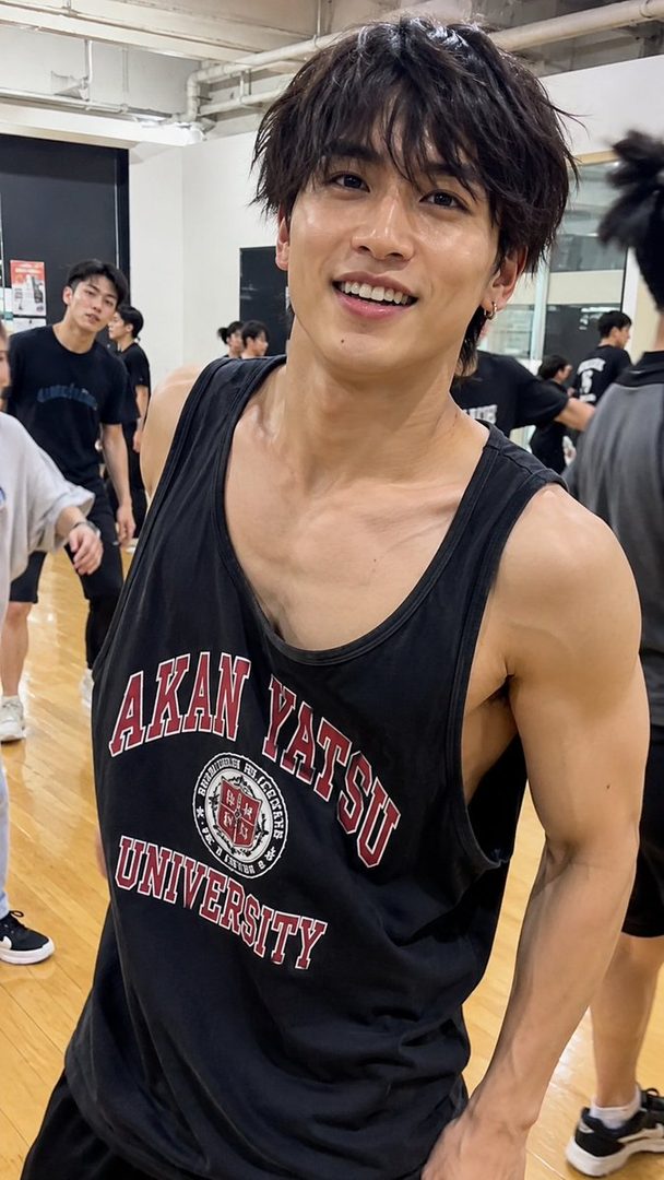 A young East Asian man with dark, messy hair smiles broadly, wearing a black tank top with 'AKAN YATSU UNIVERSITY' printed on it, in an indoor gymnasium.
