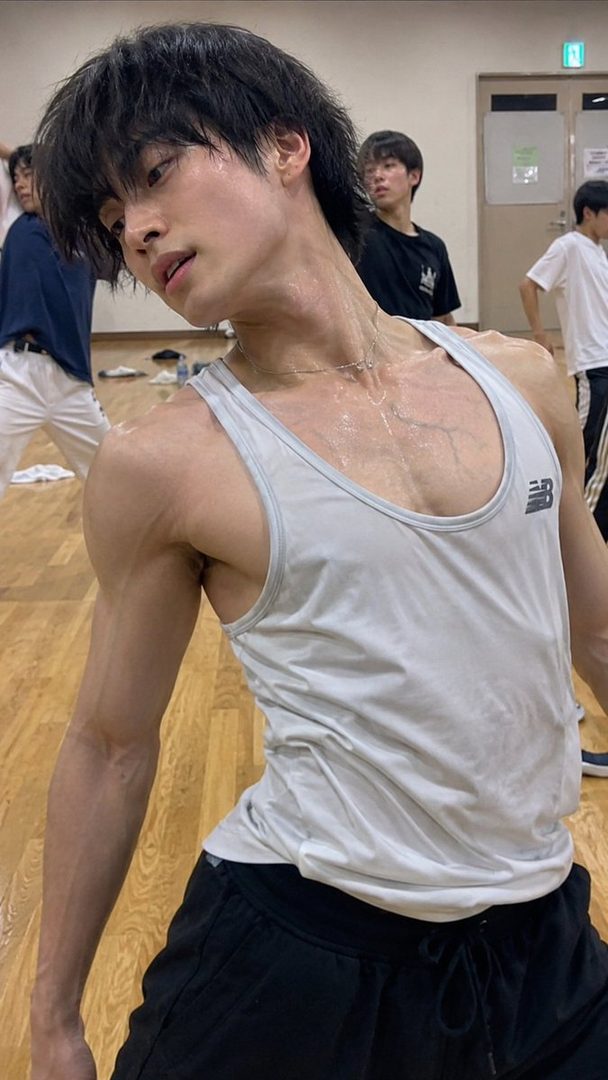 A young man with black hair, wearing a white tank top and black shorts, sweats during a dance practice session in a studio.