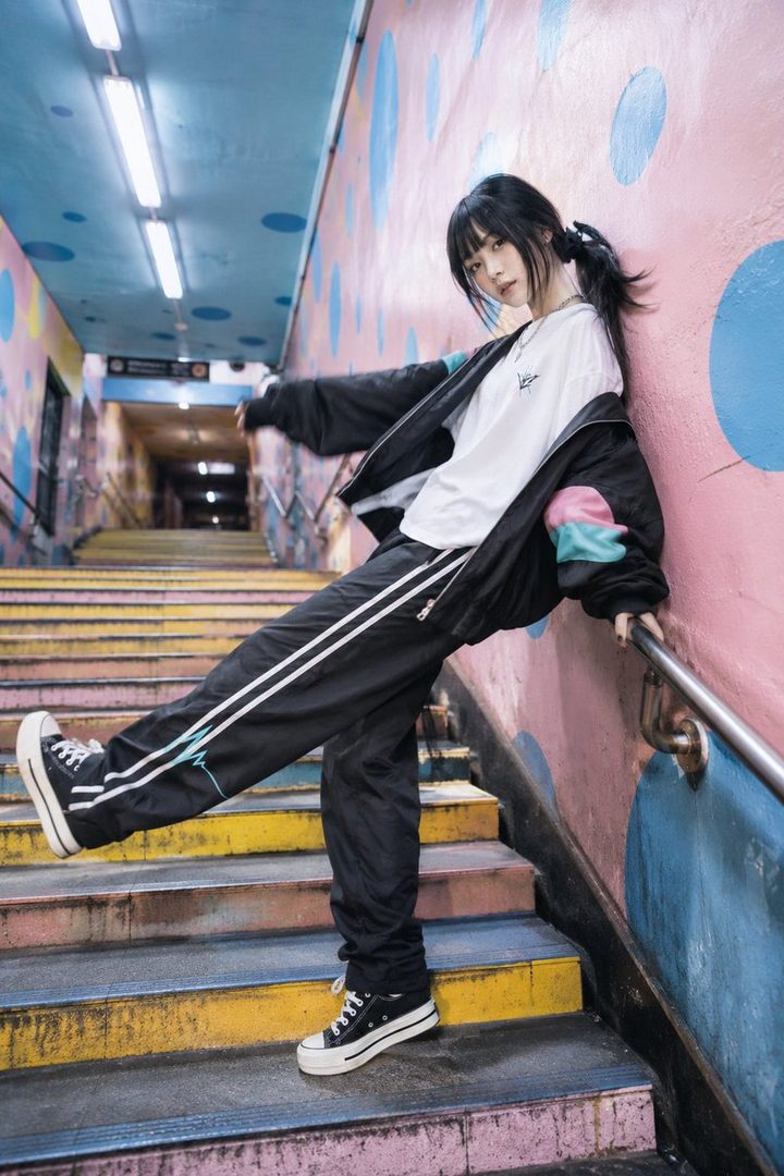 A young woman with black hair in a ponytail poses on colorful stairs, wearing a black jacket over a white t-shirt and black track pants.