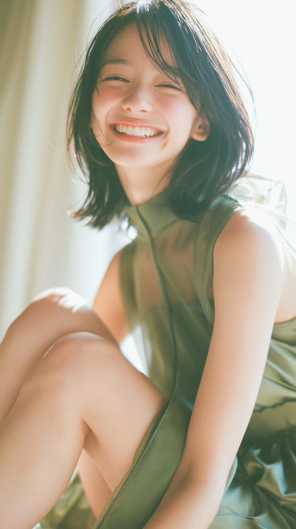 A young woman with short black hair smiles brightly, sitting indoors with soft light illuminating her face and green dress.