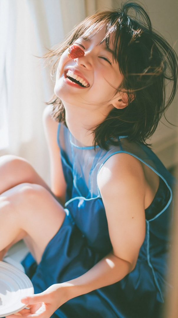 A young woman with short dark hair laughs heartily, with a strawberry placed over her eye, wearing a blue dress.