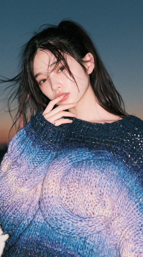 A young woman with long black hair tied in a ponytail, wearing a colorful knitted sweater, poses outdoors at dusk.
