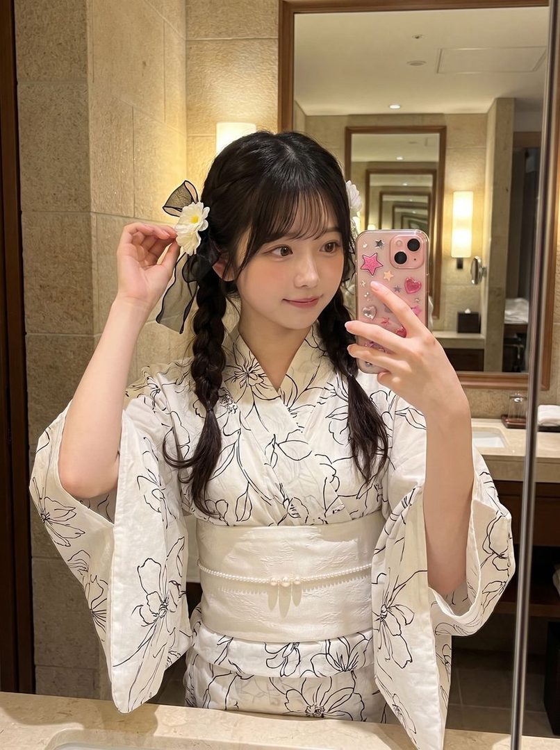 A young woman with black hair in twintails, wearing a white yukata with a floral pattern, takes a selfie in a mirror.