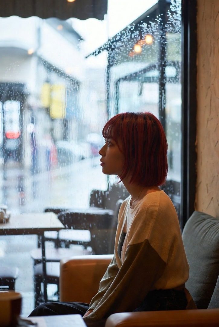 A young woman with short red hair looks out a rain-streaked window, sitting indoors.