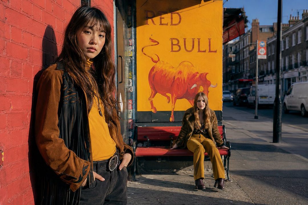 Two young women posing on a city street, one leaning against a brick wall and the other sitting on a bench in front of a Red Bull advertisement.
