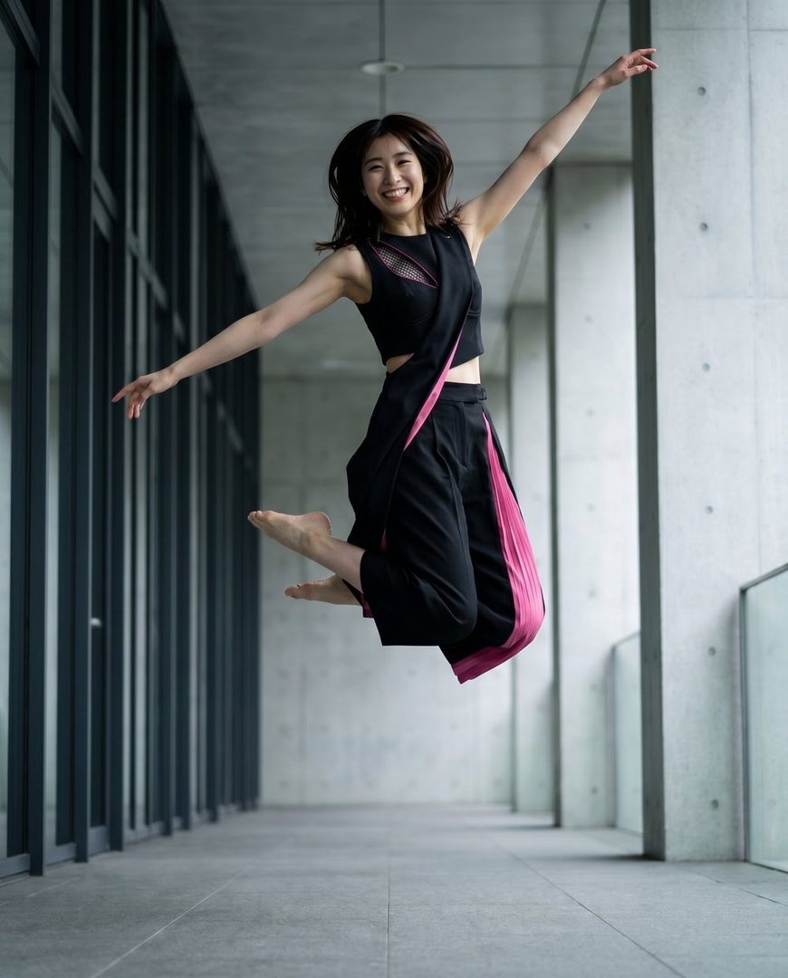 A young woman with dark hair jumps in the air, wearing a stylish black and pink outfit, with a modern architectural background.