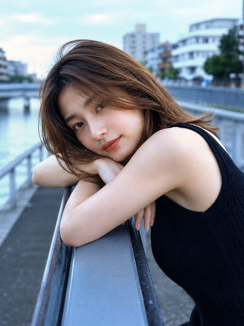 A young woman with long brown hair leans on a railing outdoors, looking thoughtfully at the camera.