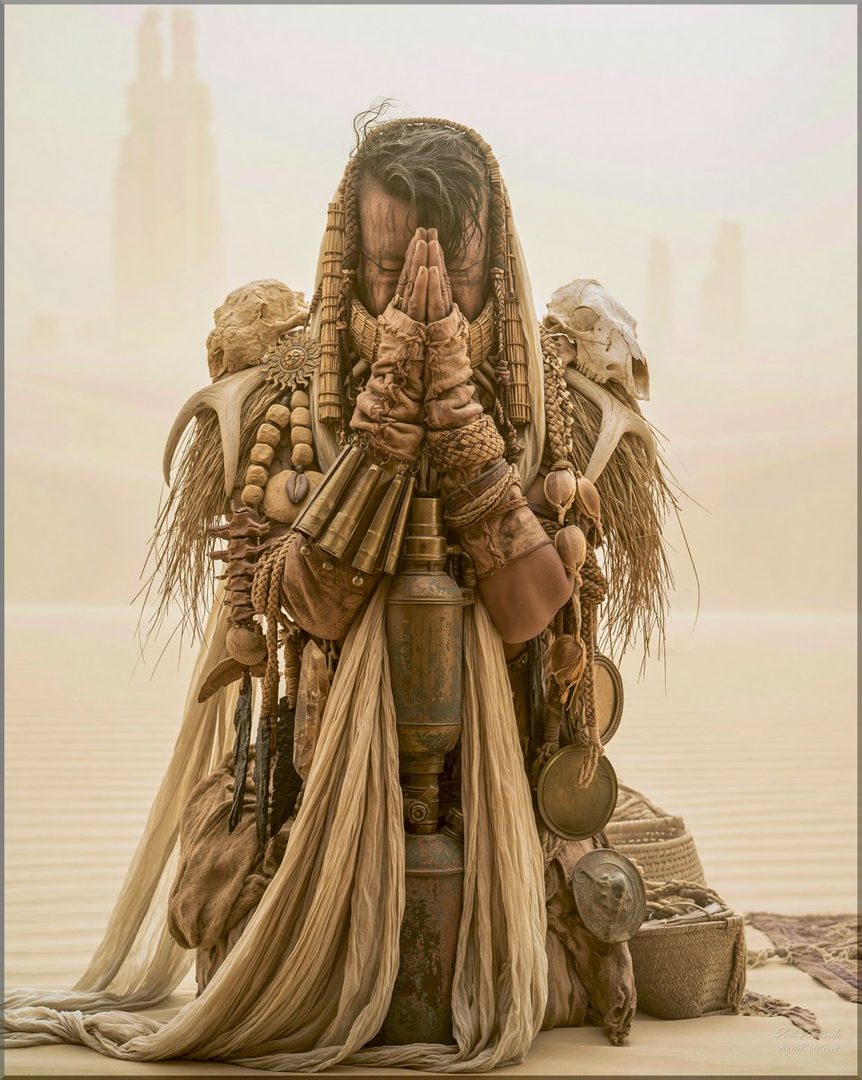 A person in elaborate desert-themed attire, with prayer beads, skulls, and fabric wraps, kneels with hands clasped in prayer in a sandy desert landscape.