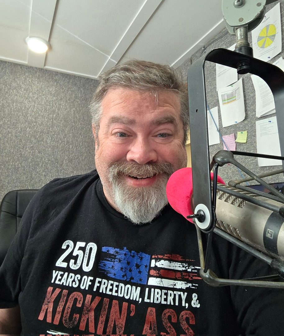 A middle-aged man with grey hair and a beard smiles at the camera in a recording studio, wearing a t-shirt with an American flag design.