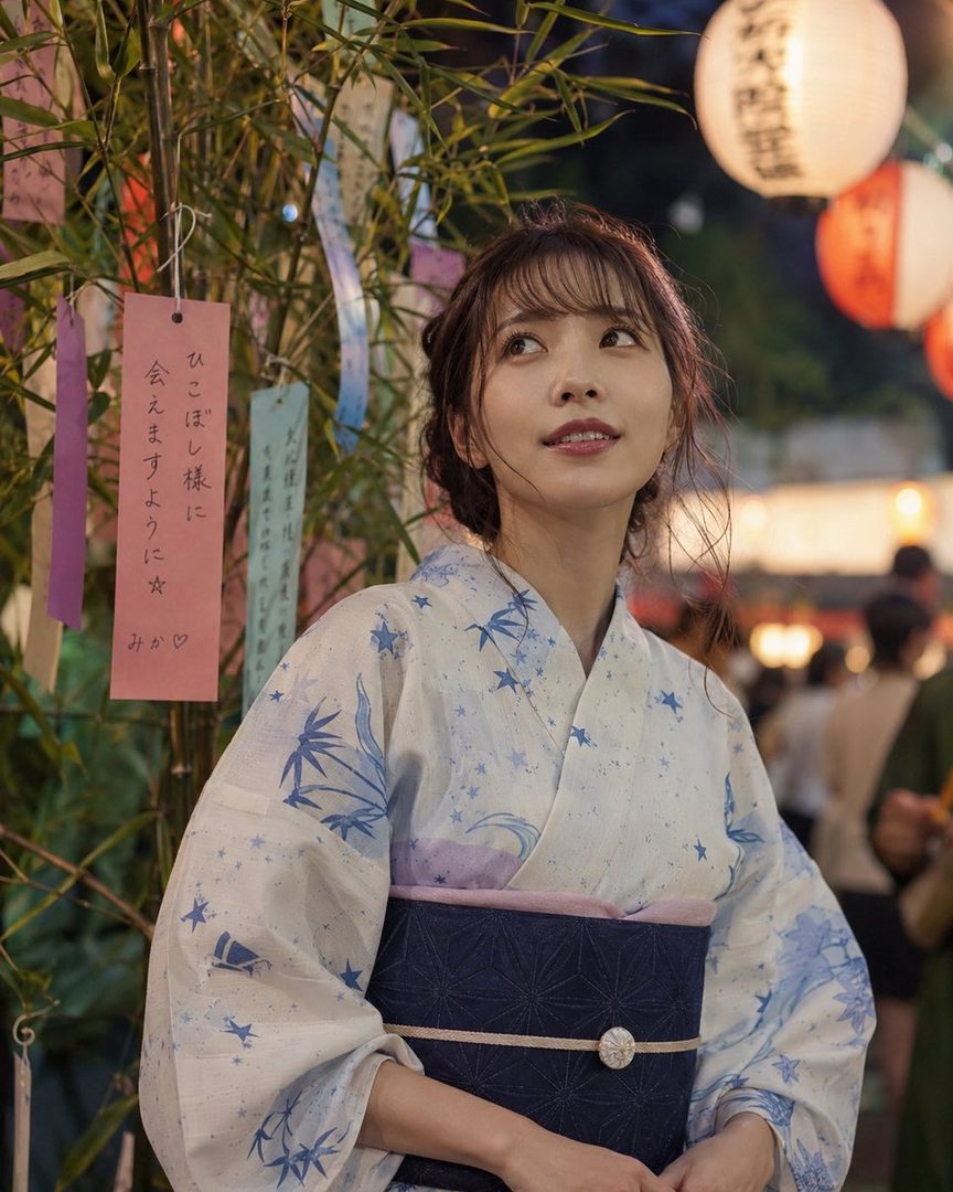 A young Japanese woman in a blue and white yukata stands outdoors at night, with bamboo and lanterns in the background.
