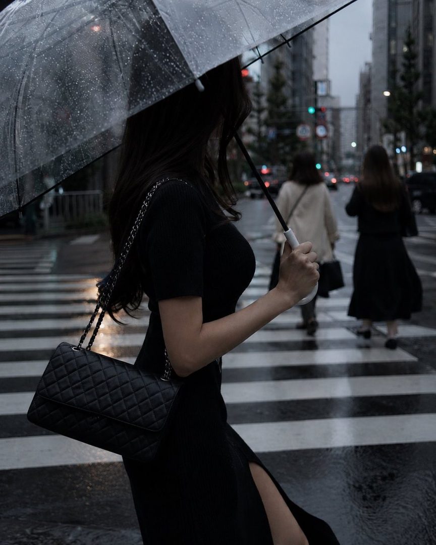A woman in a black dress walks across a wet city street at night, holding a clear umbrella and a black handbag.