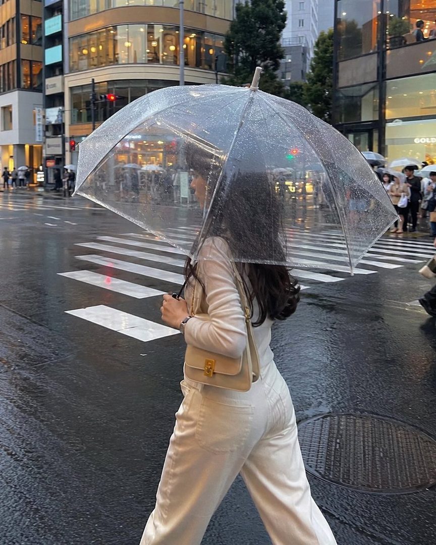 A woman walks across a wet city street in the rain, holding a clear umbrella and looking at her phone.