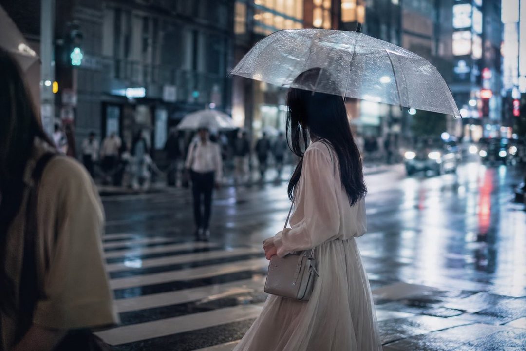 A woman walks across a wet city street at night under a clear umbrella, with blurred city lights and traffic in the background.