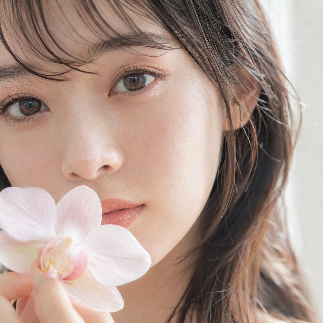 A close-up portrait of a young East Asian woman with long brown hair, holding a delicate pink orchid near her lips, with soft lighting.