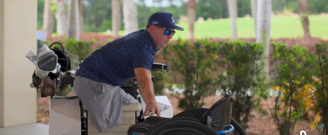 A man in a blue shirt, cap, and sunglasses is seen on a golf course, leaning towards a golf cart with clubs.
