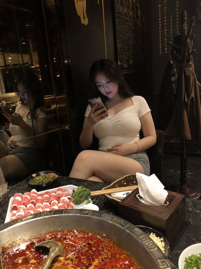 A young East Asian woman in a white top and grey shorts sits at a hotpot restaurant, looking at her phone.
