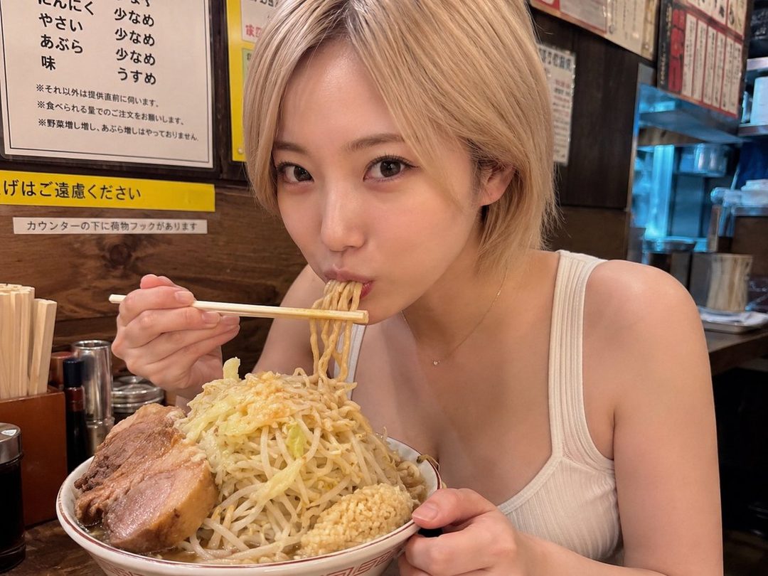 A young woman with short blonde hair eating a large bowl of ramen in a restaurant.
