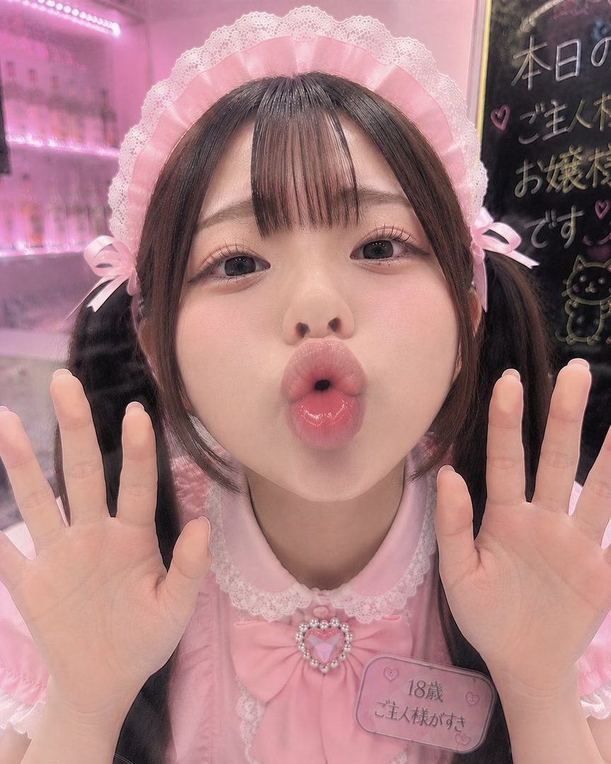 A young woman with dark hair in pigtails and bangs, wearing a pink maid outfit and a lace headband, makes a kissing face towards the camera.